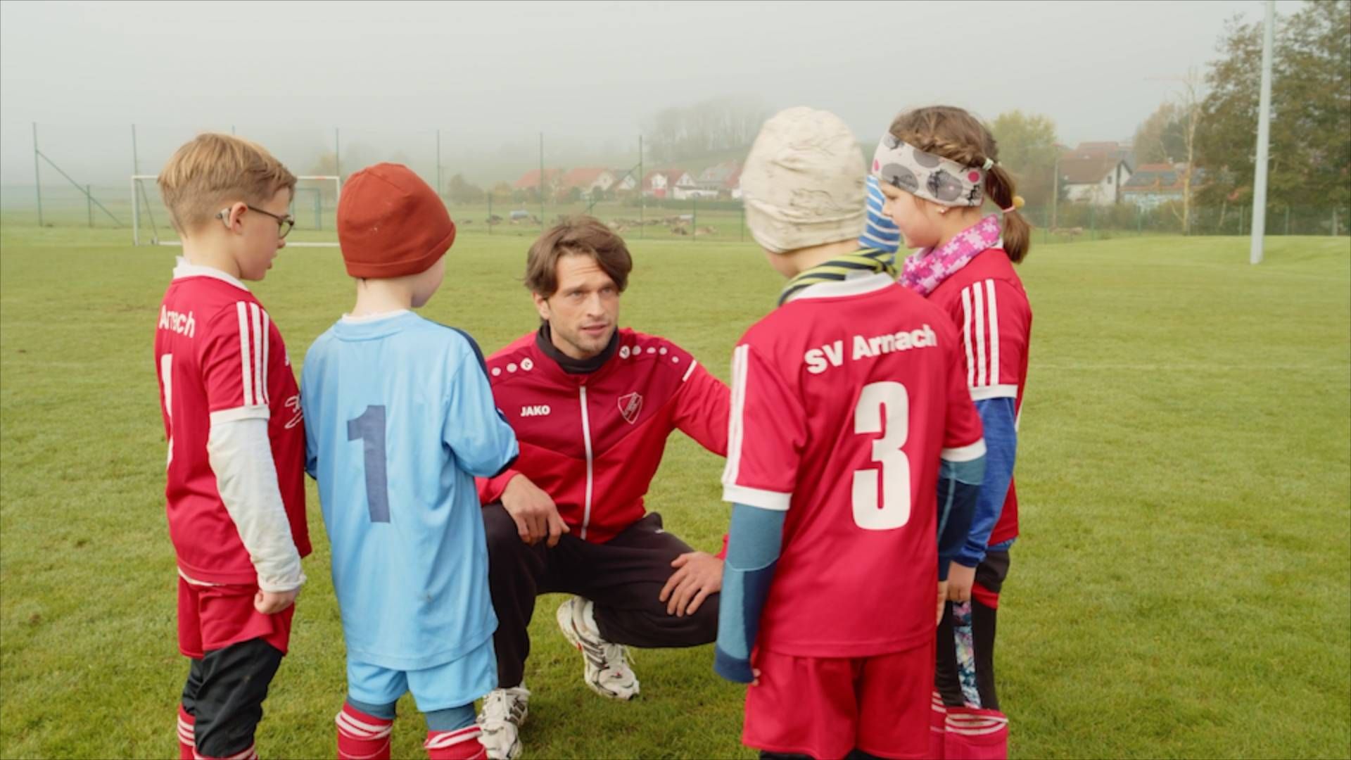 Thumbnail  - Ein junger Fußballtrainer bespricht sich kniend mit ein paar Kindern seines Kinder-Fußballteams.