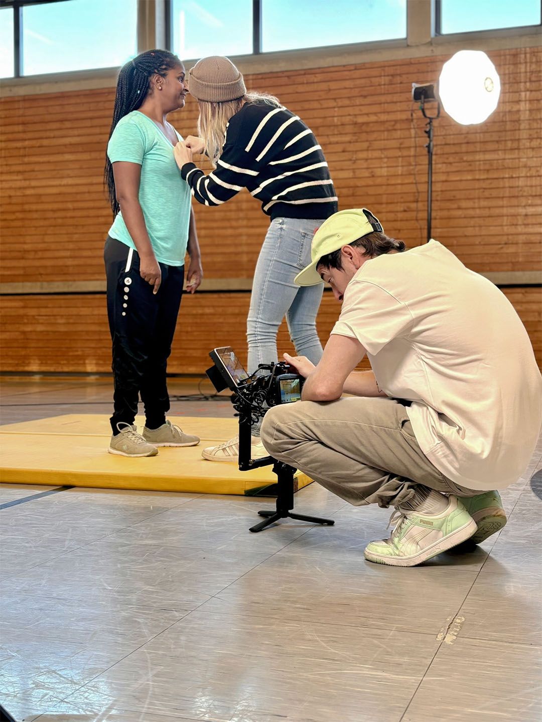 Filmteam bei der Arbeit in einer Turnhalle. Ein Mikrofon wird an einer Schauspielerin befestigt.