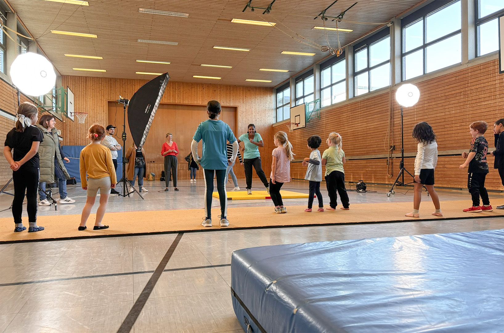Kinder am Turnen in einer Turnhalle. Im Hintergrund ist eine Filmcrew mit Lampen und weiterer Ausrüstung zu sehen.