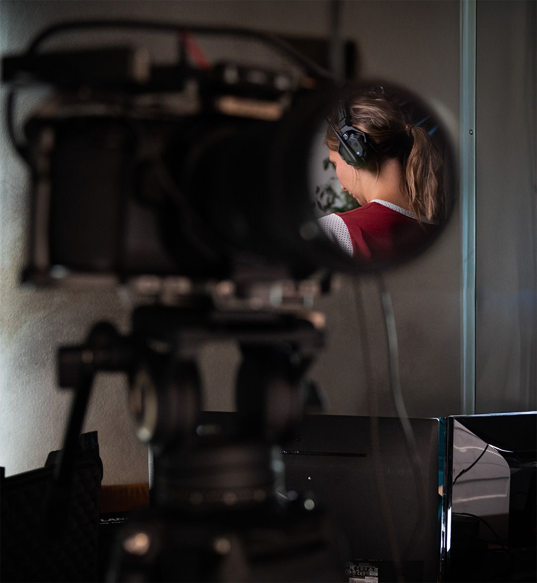 Blick auf eine professionelle Kamera. Eine Person mit Kopfhörern, während einer Video- oder Audioaufnahme, wird in der Linse gespiegelt.